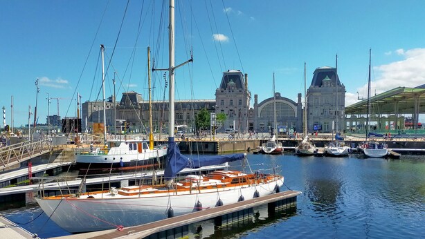 Un port de plaisance où sont amarrés plusieurs bateaux, un grand bâtiment avec un clocher en arrière-plan, et une promenade couverte.