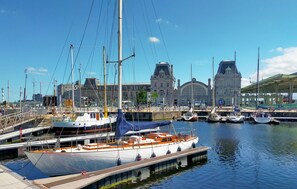 Un port de plaisance où sont amarrés plusieurs bateaux, un grand bâtiment avec un clocher en arrière-plan, et une promenade couverte.