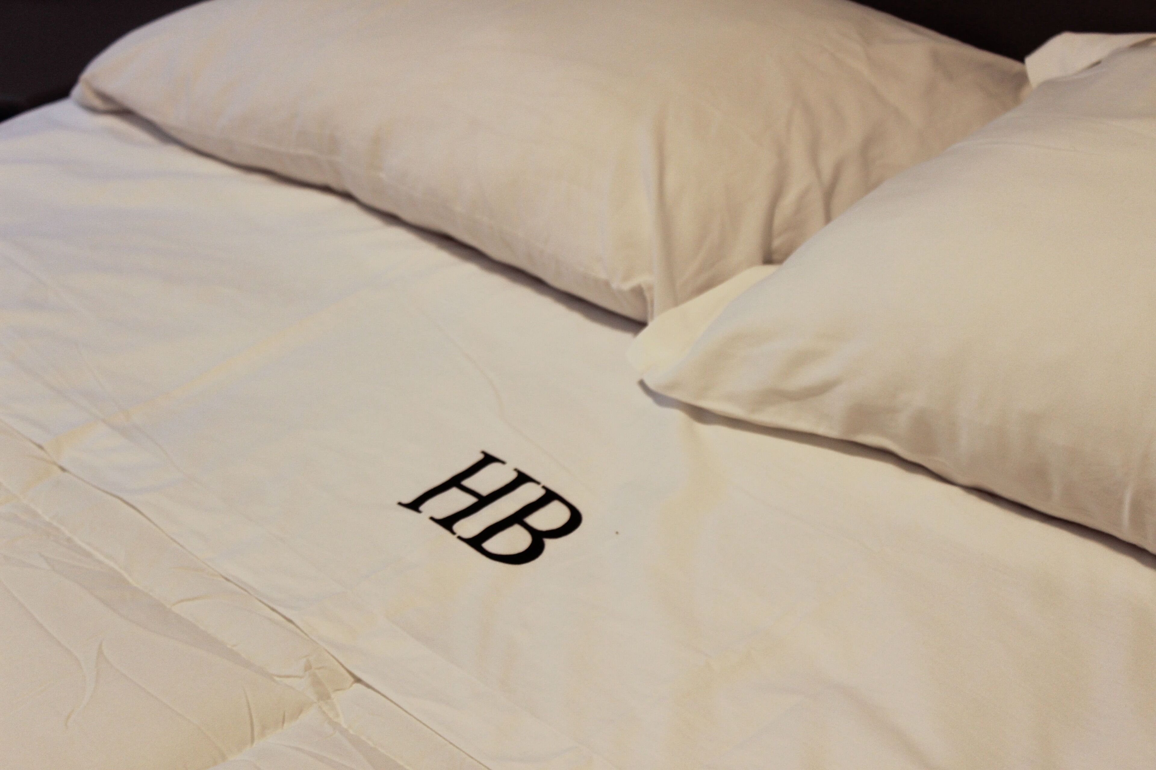Una cama con sábana blanca y dos almohadas, una con el logo "HB" visible.