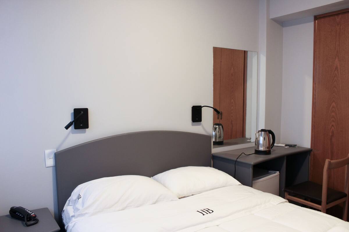 A hotel room with a bed, bedside table, a desk with a chair, a kettle, and a mirror.