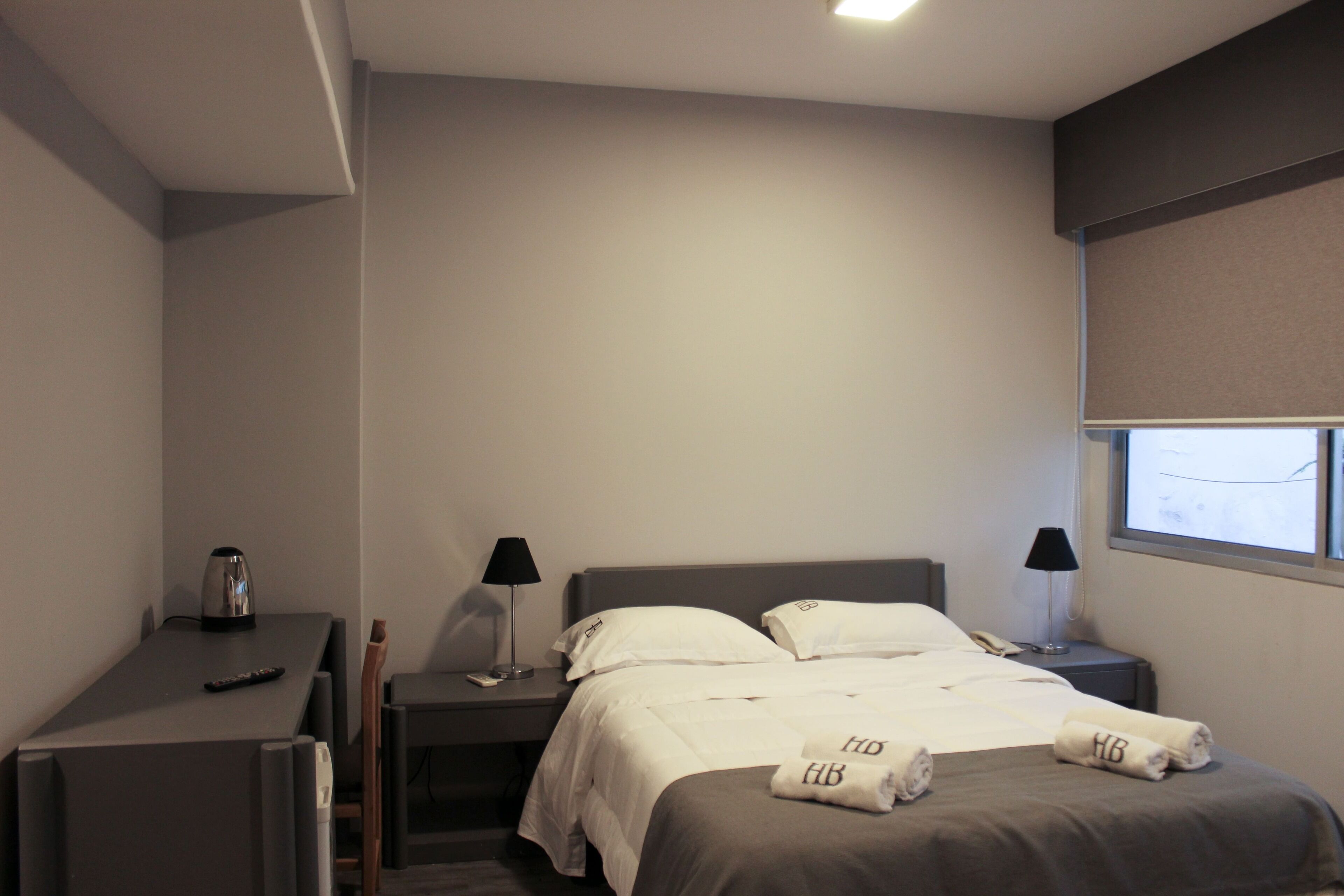 A hotel room with a bed, bedside tables, a desk, and a window with blinds.