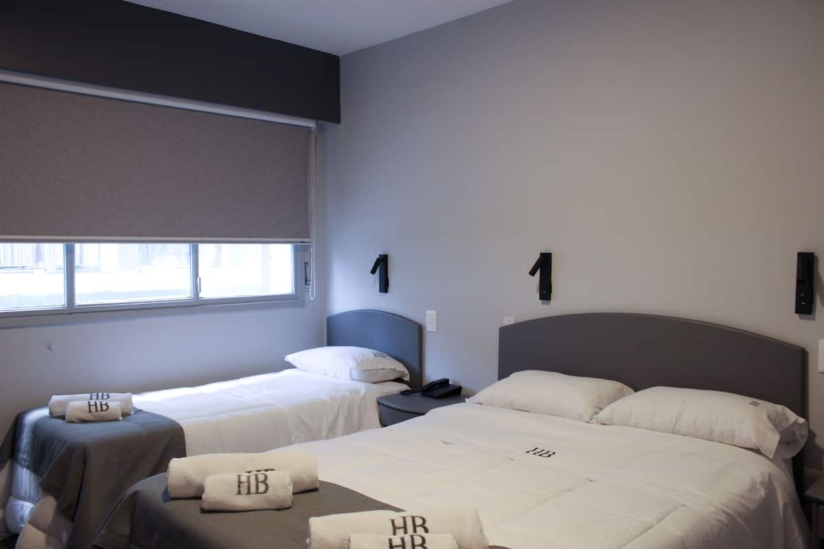 A hotel room with two beds, each with a white bedspread and pillows, a bedside table with a lamp, and a window with blinds.