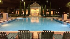 Ein Schwimmbecken im Vordergrund mit Liegestühlen, im Hintergrund ein Gebäude mit einem Dreiecksdach und ein gut beleuchteter Eingang.
