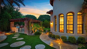 Un jardín bien cuidado con un sendero de piedra que lleva a una casa con ventanas arqueadas y una pérgola cubierta de plantas.