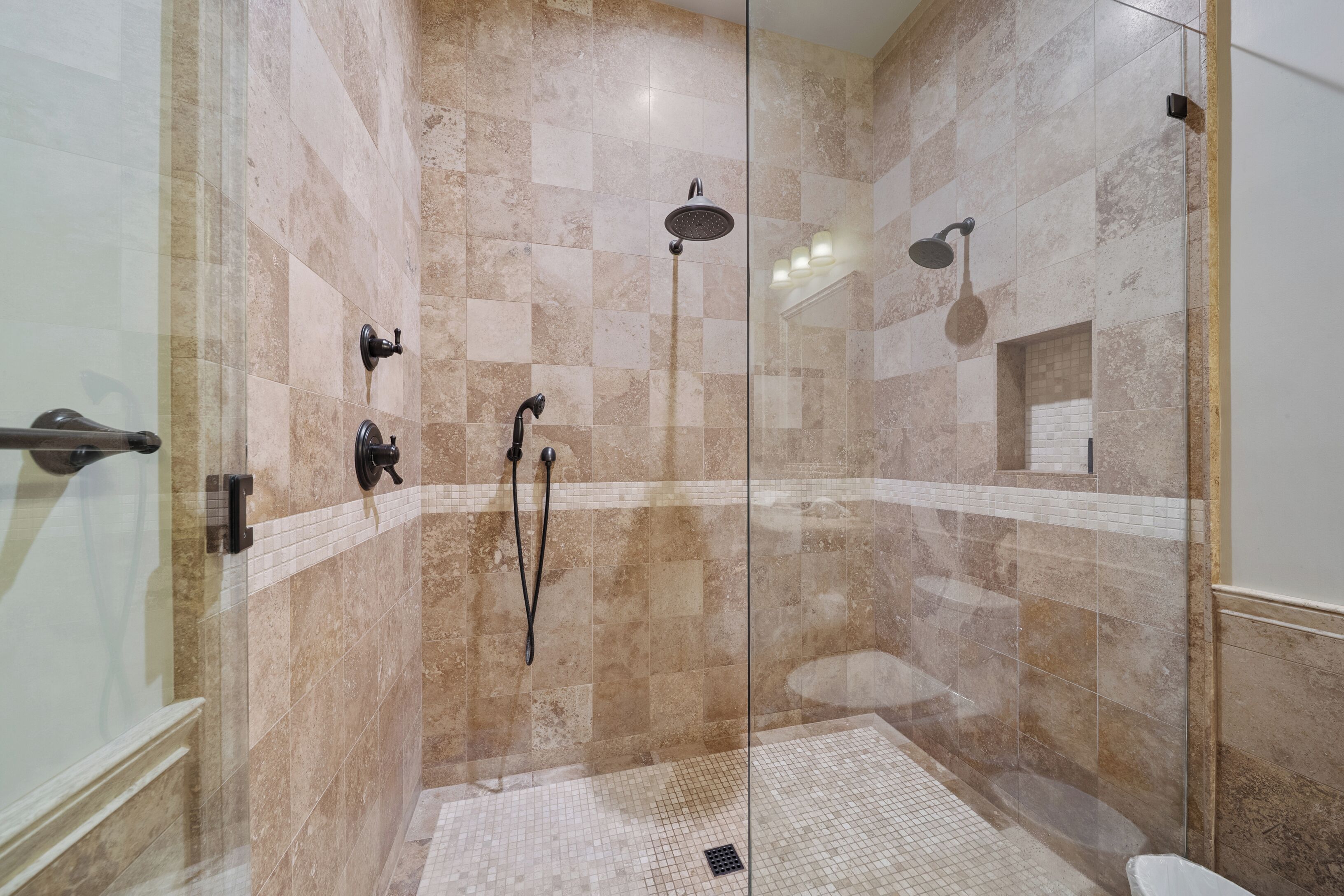 A glass-enclosed shower with multiple showerheads and a tiled floor.