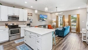 A modern kitchen with white cabinetry, stainless steel appliances, and a central island. There is a dining area with a blue sofa and orange chairs. - Pet Friendly East Hill New Build - Walk to Graffiti Bridge - Close to Downtown and Pensacola Beach (Pensacola)