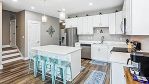 A modern kitchen with white cabinets, a stainless steel refrigerator, and a coffee machine. There are two bar stools and a large island. - Pet Friendly East Hill New Build - Walk to Graffiti Bridge - Close to Downtown and Pensacola Beach (Pensacola)