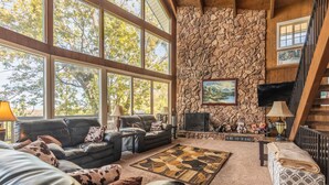 Una sala de estar acogedora con chimenea de piedra, muebles de cuero y grandes ventanales con vistas a árboles.
