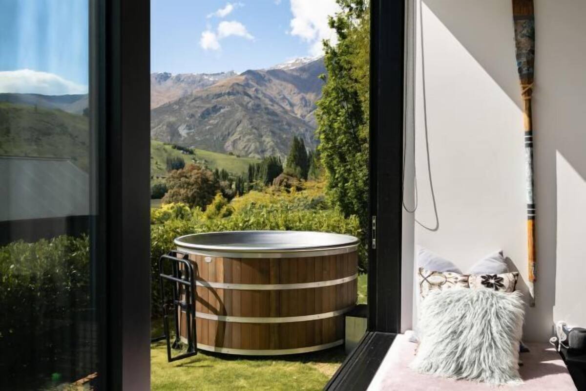 Een kamer met een groot raam met uitzicht op een bergachtig landschap, een houten jacuzzi en een gezellig bed met zachte kussens.