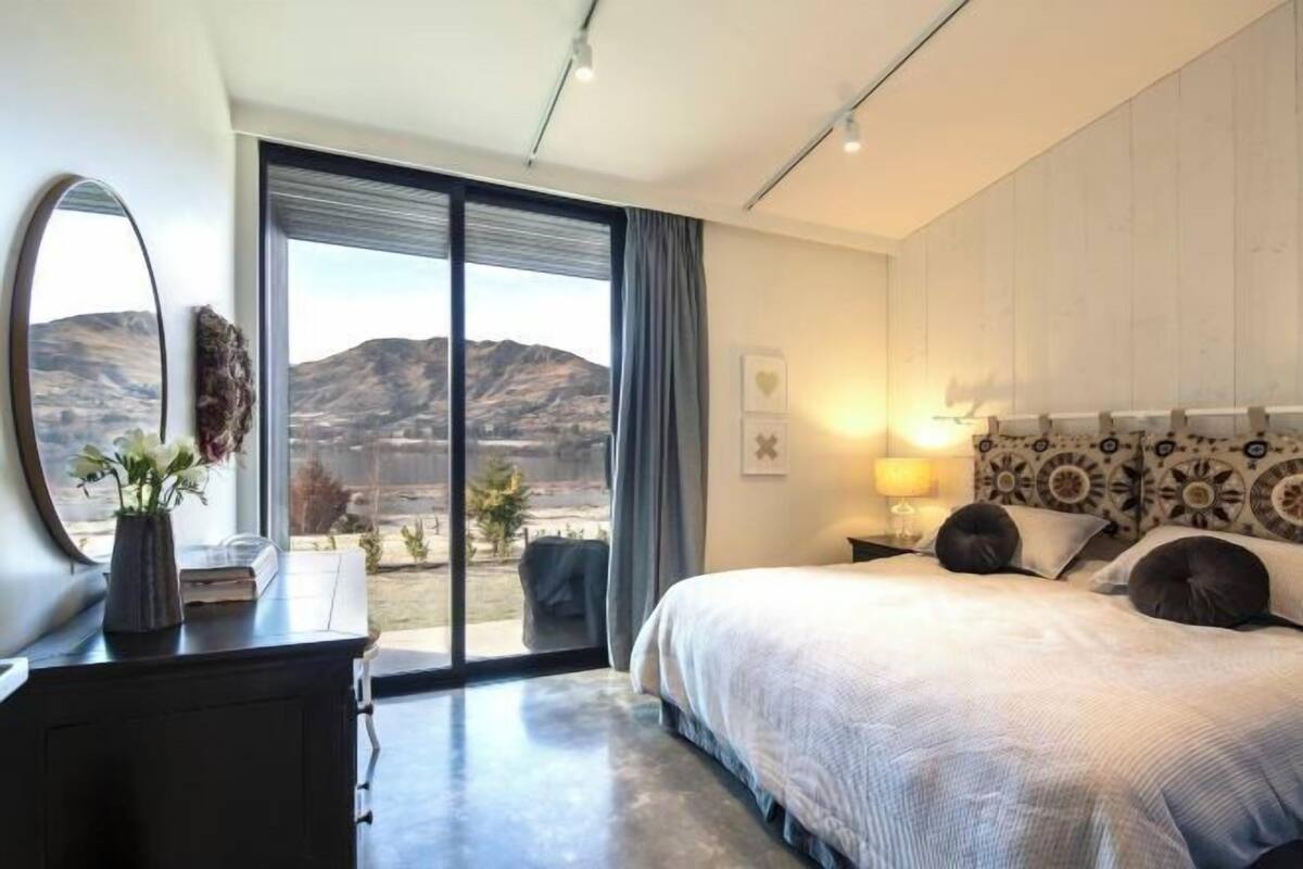 Een slaapkamer met een groot bed, een bureau met spiegel, uitzicht op bergen door een schuifdeur, en een hoofdbord met patroon.