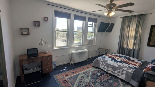 Un dormitorio con una cama, un escritorio con una computadora portátil, un radiador, un ventilador de techo y una ventana con vista a la calle.