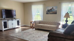 A living room with a flat-screen TV on a cabinet, a beige sofa, a dark leather armchair, a patterned rug, and a painting on the wall. -  Farmhouse close to wedding venues, hunting plantations in Whigham - 3 bedrooms (Whigham)