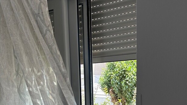 Quarto com uma janela grande, cortina e vista para uma varanda com plantas e flores.