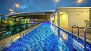 A rooftop swimming pool with a glass barrier, metal railings, and a view of the cityscape at dusk.