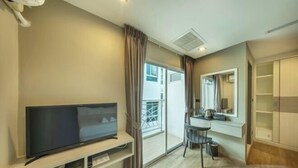 A modern hotel room with a flat-screen TV on a wooden stand, a small desk with a chair, a large mirror, and a sliding glass door leading to a balcony.