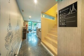 A hotel lobby with a staircase, a wall with a cityscape mural, and a sign with room numbers. - Ralph Poshtel Pratunam (Bangkok)