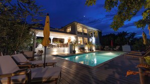 Un espace aménagé au bord de la piscine, avec des chaises longues, une terrasse en bois et une maison à plusieurs niveaux, dotée d’un éclairage extérieur.