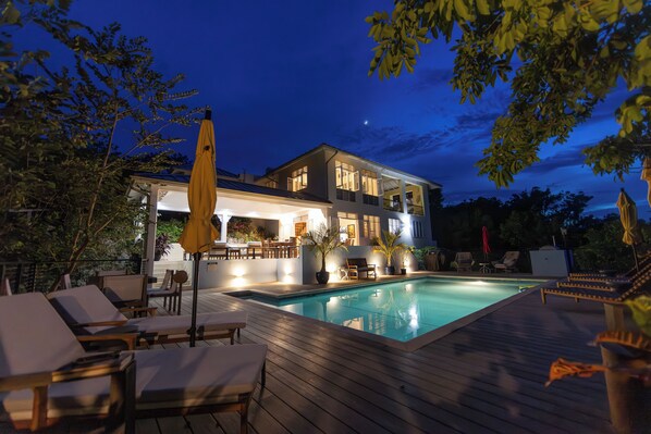 A poolside area with lounge chairs, a wooden deck, and a house with multiple levels and outdoor lighting.