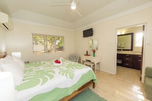 Un dormitorio con cama, escritorio, silla, televisor y baño visible a través de una puerta abierta.