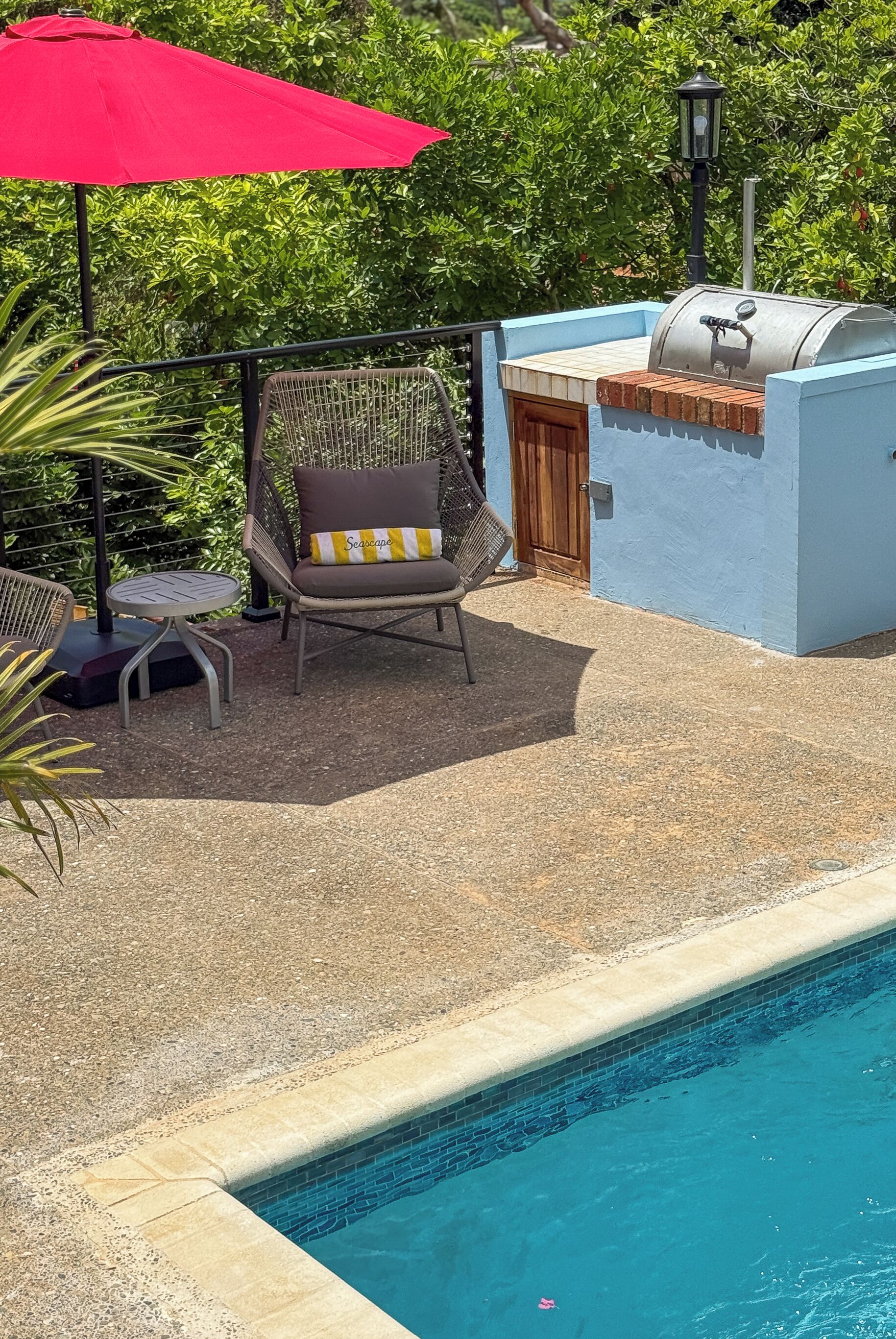 Ein Poolbereich mit Grill, einem Stuhl mit Sitzkissen und einem roten Sonnenschirm.