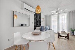 Una sala moderna con una mesa redonda blanca, sillas blancas, un ventilador de techo y un ventanal con cortinas translúcidas.