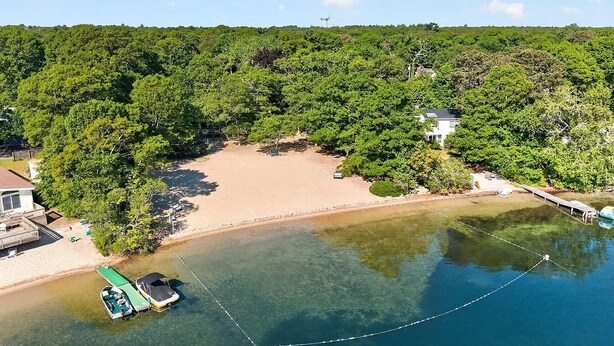 Vista aérea de uma praia com um cais para barcos, uma casa e floresta densa.