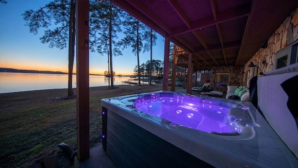 Un bain à remous avec vue sur un lac au crépuscule, un patio couvert et un mur en pierre.