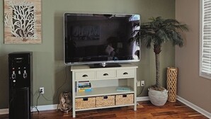 A living room with a flat-screen TV on a white stand, a small black speaker, a wooden floor, a potted plant, and a wall-mounted artwork. - 3-bedroom townhome in beautiful North Wildwood with AC two blocks from the beach (North Wildwood)