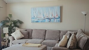 A living room with a sectional sofa, an ottoman, a ceiling fan, a painting of sailboats, and a floor lamp. - 3-bedroom townhome in beautiful North Wildwood with AC two blocks from the beach (North Wildwood)