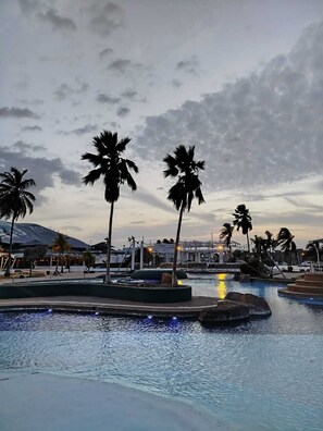 Área de piscina en un resort, con palmeras, un elemento acuático y una montaña al fondo.