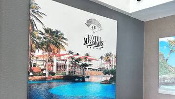Un hall d’hôtel orné d’une grande fresque murale représentant une piscine et des palmiers, avec deux fauteuils gris, une petite table d’appoint ronde en bois et un panier tressé.