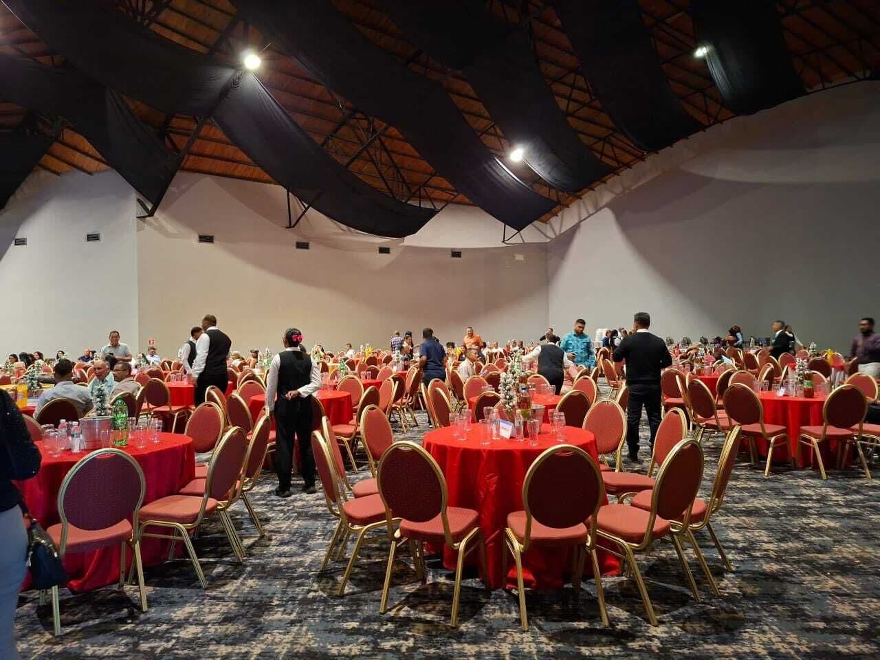 A banquet hall with round tables covered in red tablecloths and gold chairs, a high ceiling with exposed beams, and a carpeted floor.