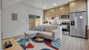 A modern kitchen with a built-in oven, microwave, and refrigerator. There is a grey sectional sofa with throw pillows, a wooden dining table, and a colorful rug.