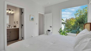 A bedroom with a large bed, a bathroom with a vanity, and a view of trees and a house through a window.