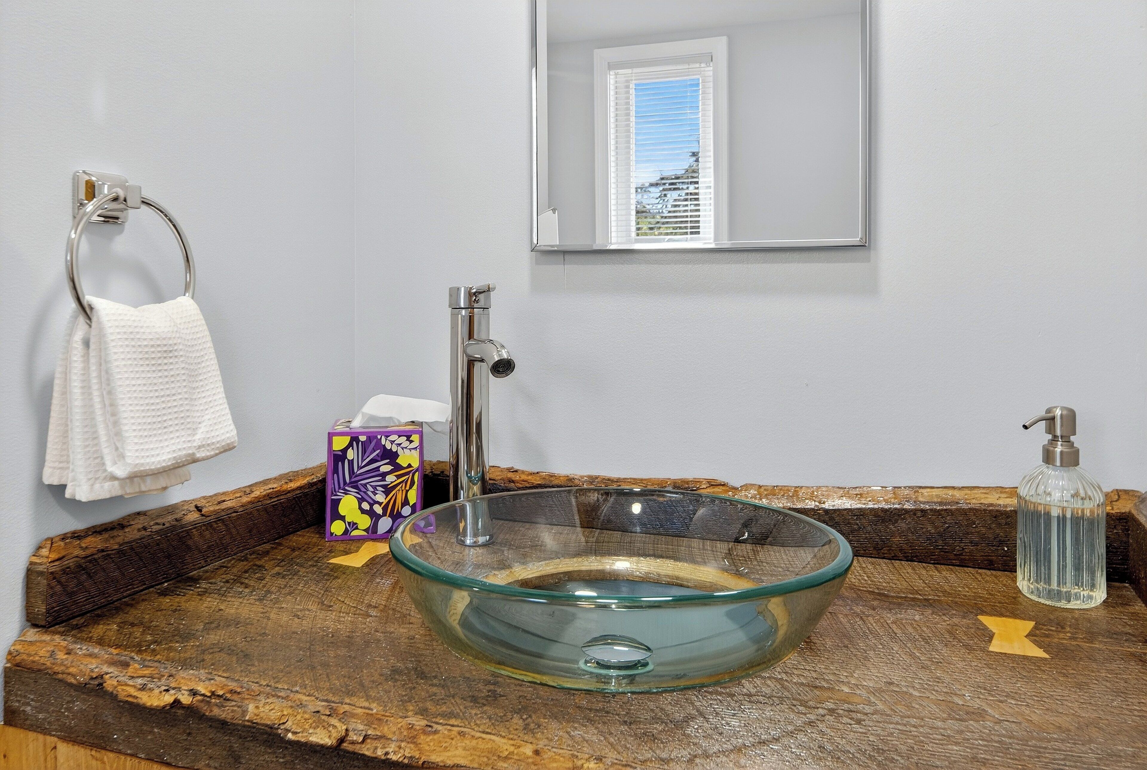 A bathroom with a glass bowl sink, a towel rack, a soap dispenser, and a mirror.