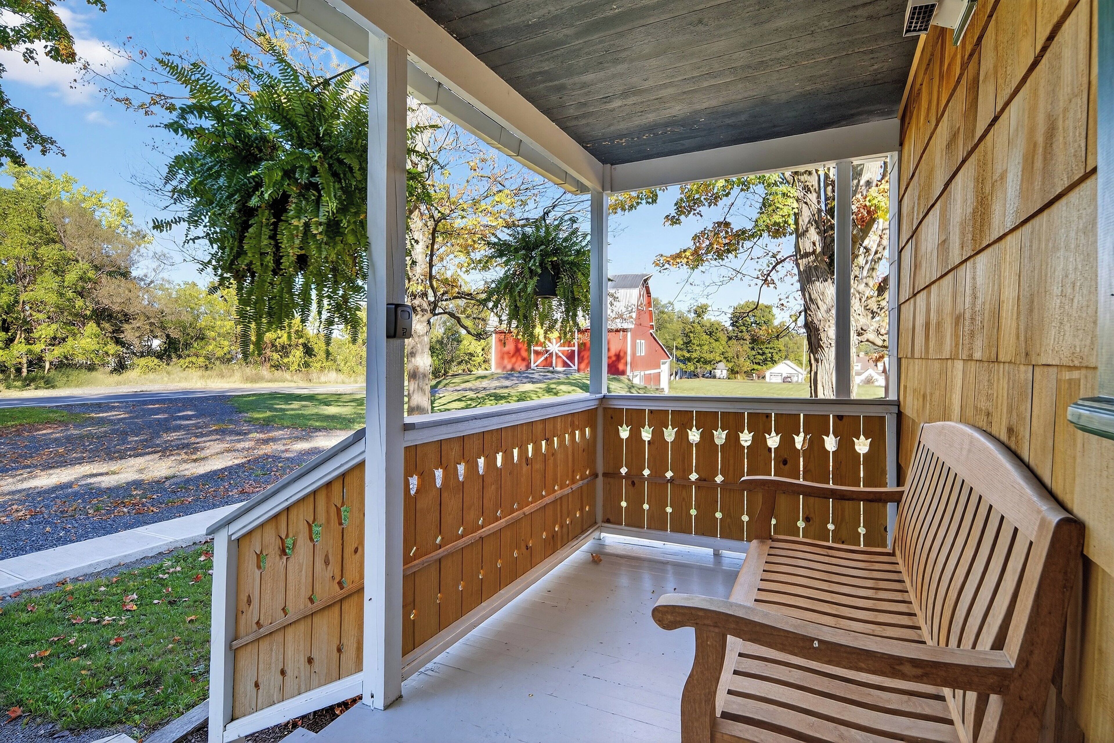 A covered porch with a wooden bench, a white railing, and a view of a red barn and trees.