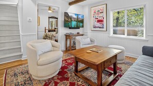Una sala de estar con una mesa de centro de madera, dos sillones blancos, un tapete rojo con estampado, un televisor montado en la pared y una ventana con persianas.