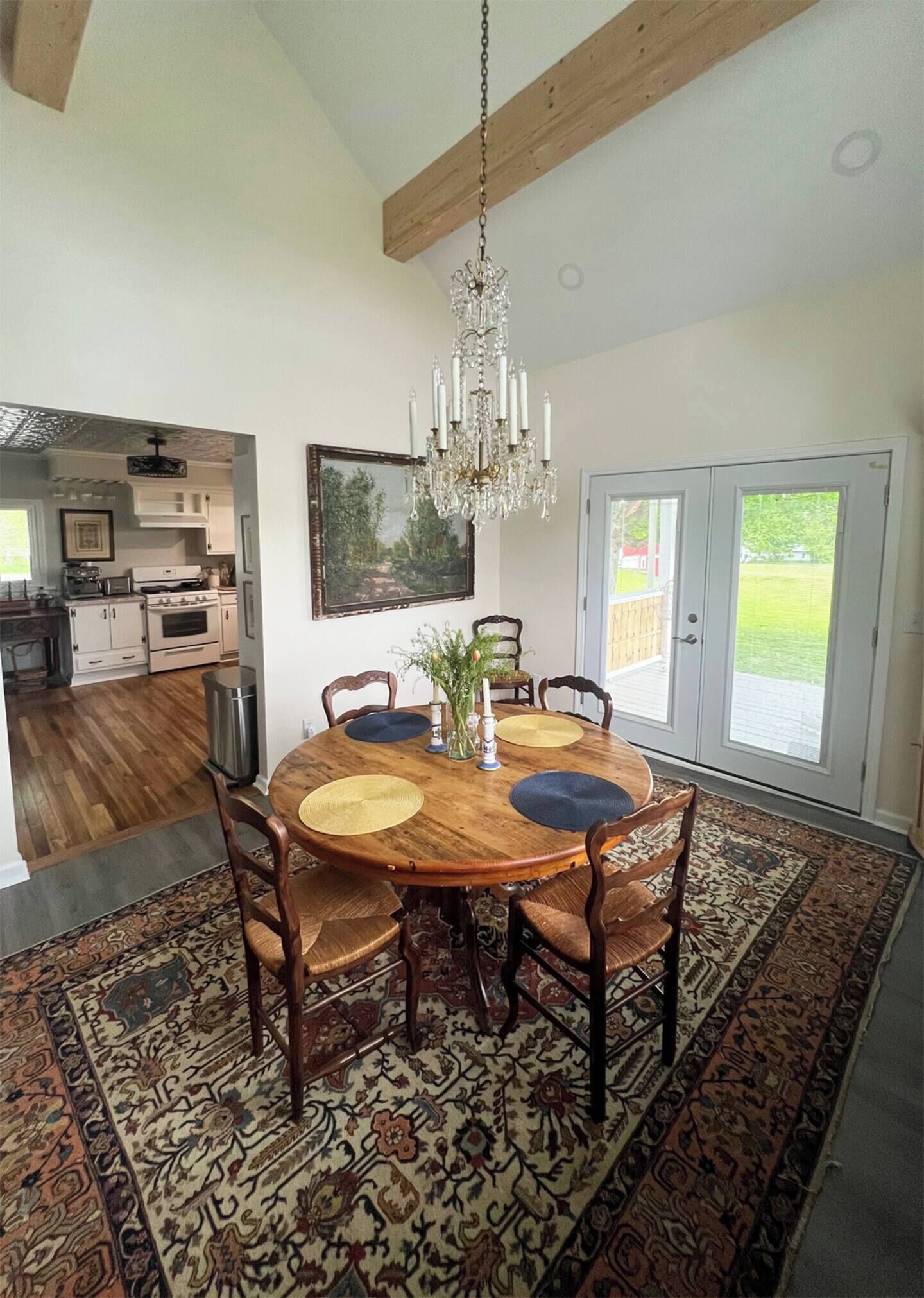 A dining area with a wooden table, chairs, and a chandelier. There is a patterned rug on the floor, and a view of the kitchen and outside through a glass door.