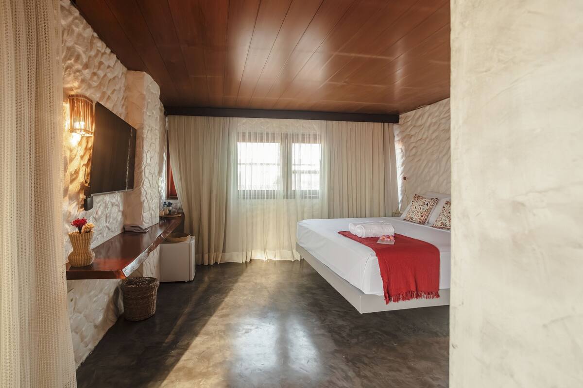 Un dormitorio con techo de madera, una cama con manta roja, una mesita de noche, una lámpara y una ventana con cortinas traslúcidas.