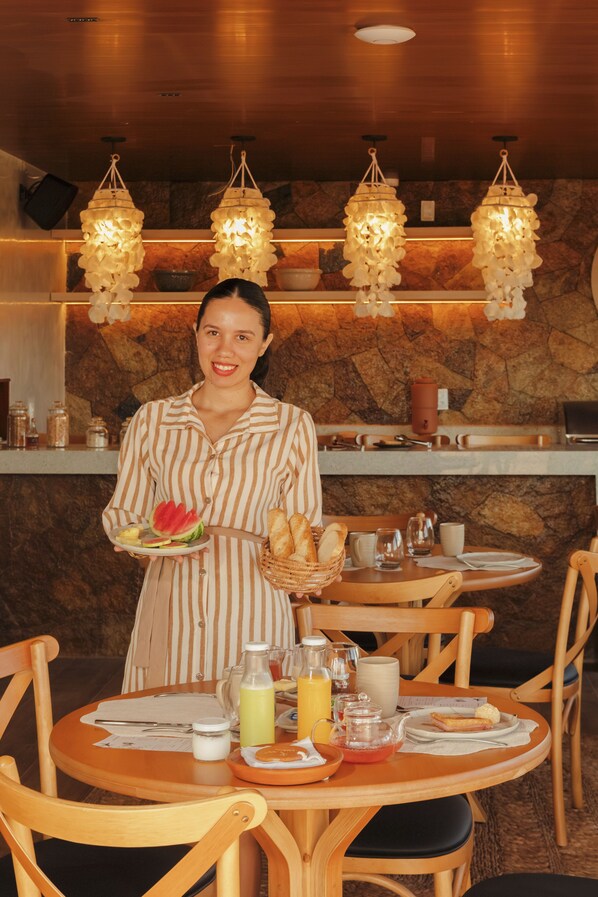 A breakfast setting with a person holding a plate of food, a basket of bread, and a glass of juice. There are wooden chairs and tables, and a stone wall in the background. - Amana Icaraizinho (Amontada)