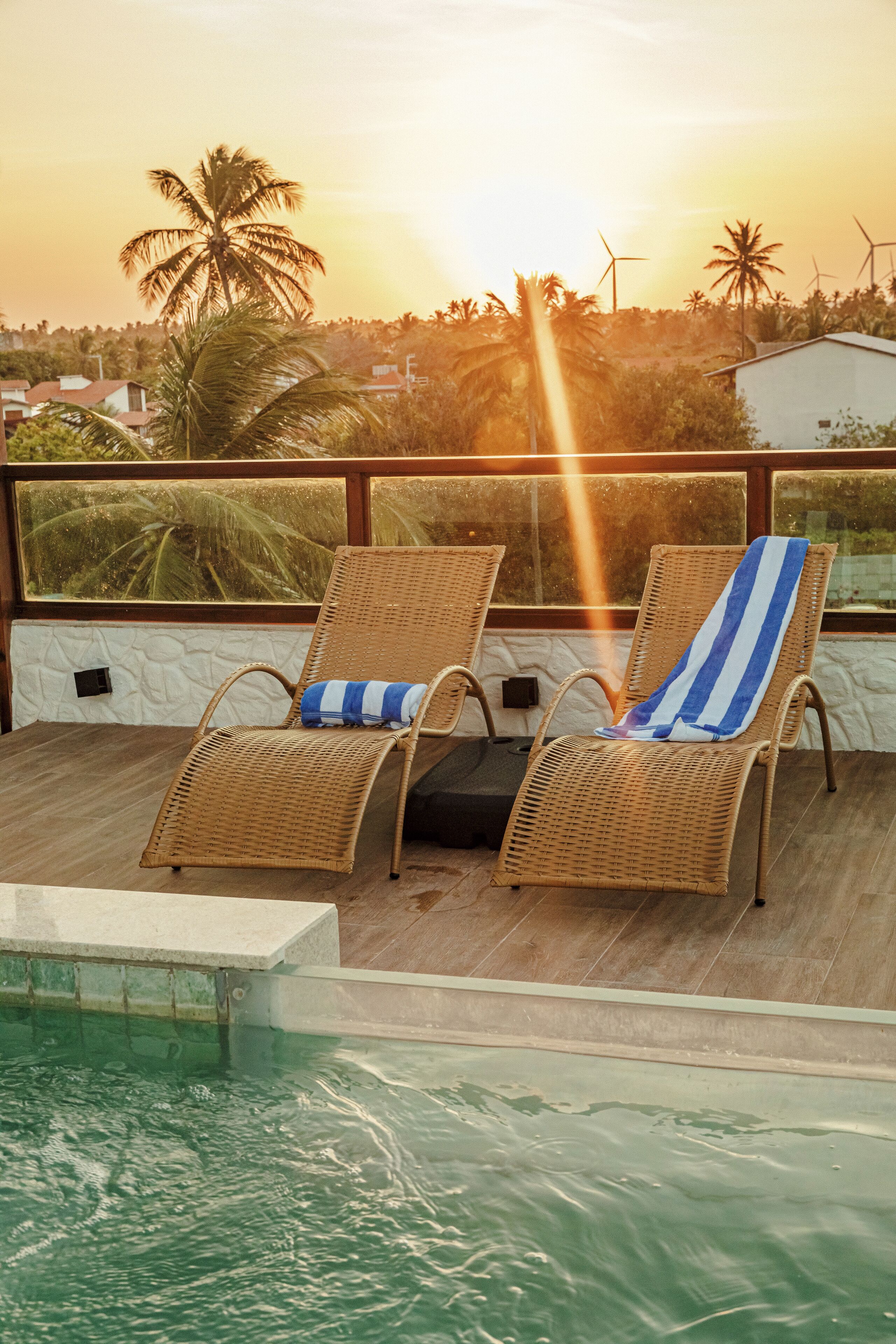 Duas poltronas de vime com toalhas listradas junto a uma piscina, com vista para um pôr do sol com palmeiras e turbinas eólicas ao fundo.