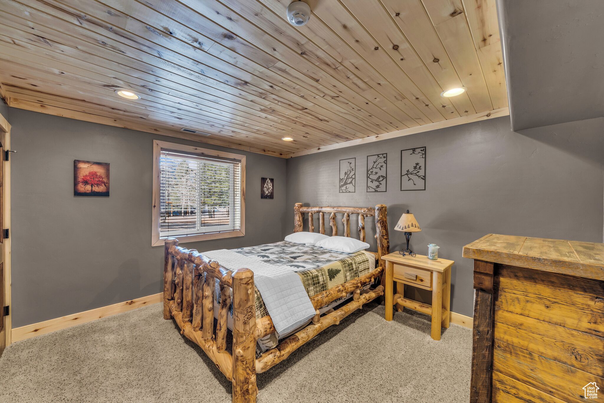 Une chambre avec un lit en bois, une fenêtre avec des stores, une table de chevet avec une lampe et un mur gris orné de tableaux encadrés.