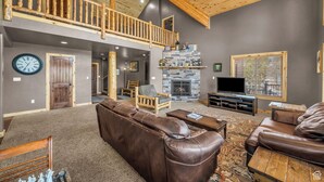 A spacious living room with a high ceiling, a stone fireplace, a wooden balcony, and a large television. - The Lost Creek Lodge - A Perfect Escape for Families, Reunions, and Friends. (Duck Creek Village)