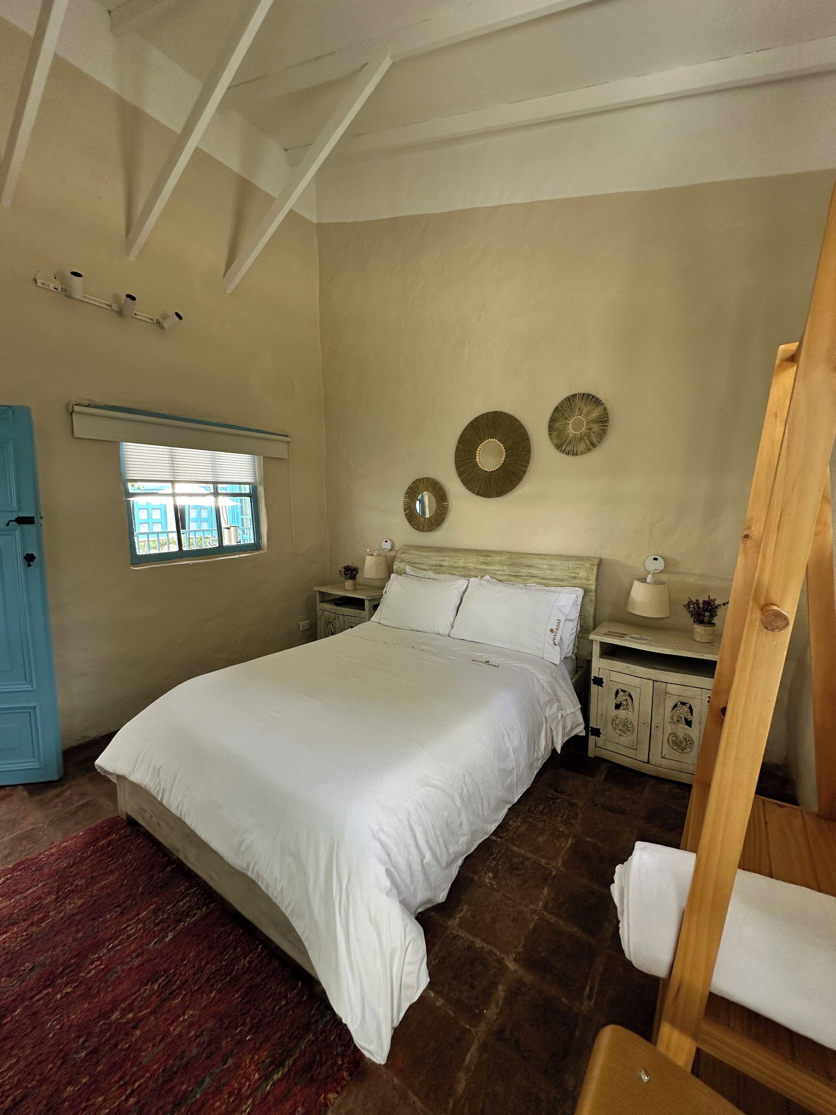 Un dormitorio con una cama grande, una silla de madera, una mesita con una lámpara y una ventana con persianas.