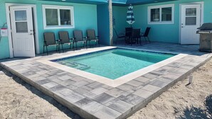 A pool area with a tiled deck, a seating area with chairs and a table, and a building with a blue exterior and white doors. - LOCATION!! LOCATION!!  SHORT WALKING TO RESTAURANTS, SHOPS, & BEACH! (Fort Myers Beach)