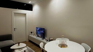 A modern living room with a round dining table, white chairs, a grey sofa, a TV, and a decorative vase. - Eliz Stay: Your Cozy Home Away From Home, Steps from the Lake. (Tiranë)
