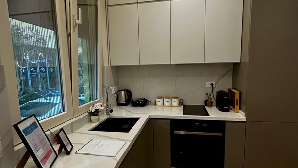 A modern kitchen with a white countertop, stainless steel appliances, and a window with a view of a garden. - Eliz Stay: Your Cozy Home Away From Home, Steps from the Lake. (Tiranë)