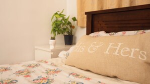A bedroom with a floral bedspread, a wooden headboard, a nightstand with a potted plant, and a pillow with "His & Hers" embroidered on it.