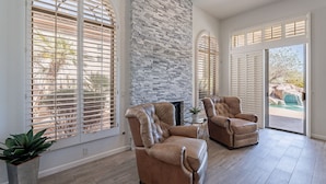 A spacious living room with a stone fireplace, two leather armchairs, and large windows with blinds.