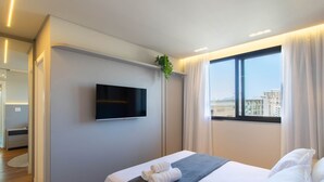 A modern hotel room with a flat-screen TV mounted on the wall, a large window with a city view, a neatly made bed with white linens and a gray blanket, and a potted plant on the windowsill. - Well-equipped Apartments 100m From the Beach - EVR (Porto Belo)